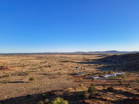An image of a small township in a remote area. There is a cluster of small light-coloured buildings and an elevated water tank in the foreground, surrounded by a large expanse of arid bushland with red dirt and rocks, grasses, trees and a bright blue sky.