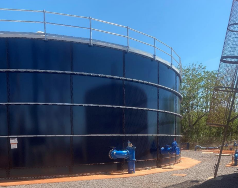 Photo of a large dark blue water tank in Galiwin’ku. There are blue valves at the bottom and a silver metal fence on top. There is pavement in the foreground and blue sky and green vegetation in the background.