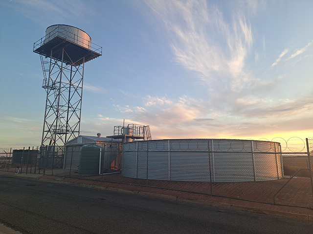 Photograph of a water treatment plant, including a water tower and circular water storage tank behind a barbed wire fence. 