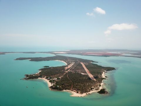 An aerial view of Gunyangara on the Gove Peninsula in north-east Arnhem Land. There is blue water and a coral reef surrounding a vegetated island. The image shows an airport runway and some roads, buildings and boats.