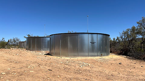 Groundwater storage tanks installed at the Iga Warta Homeland. Three large silver water storage tanks surrounded by dirt, rocks and short green shrubs. The tanks in the foreground are new.