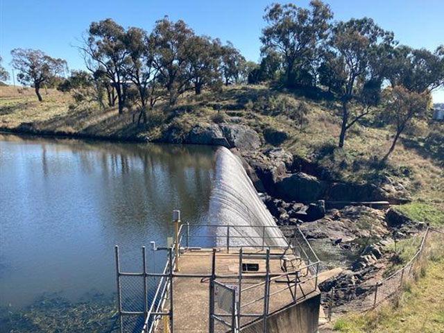 Molong Creek dam wall with water spilling over the top. A concrete path and railings lead to the edge of the dam wall. Grass and trees surround the area under a clear blue sky.