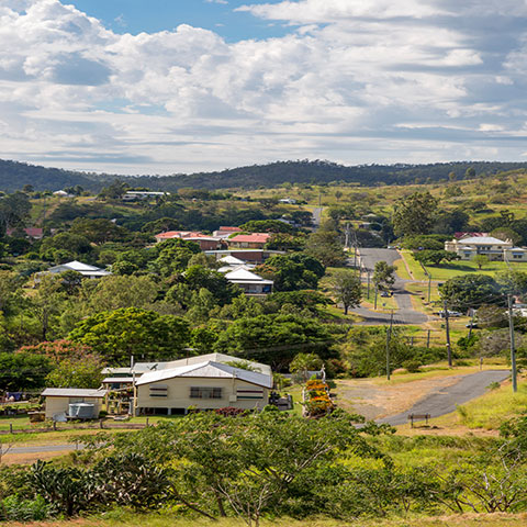 A residential street in the small rural town of Mt Morgan. There is a lot of open space and the homes are spread out with trees between and around them. There are hills in the background and clouds in the sky.