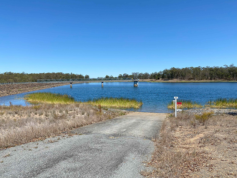 Photograph of a dam filled with water with an access road in the foreground and trees and green shrubs in the background under a blue sky.