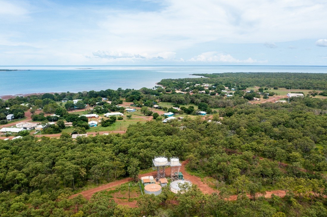 An aerial view of a small community. There are water tanks in the foreground, with a band of trees and scattered buildings behind them, leading to water.