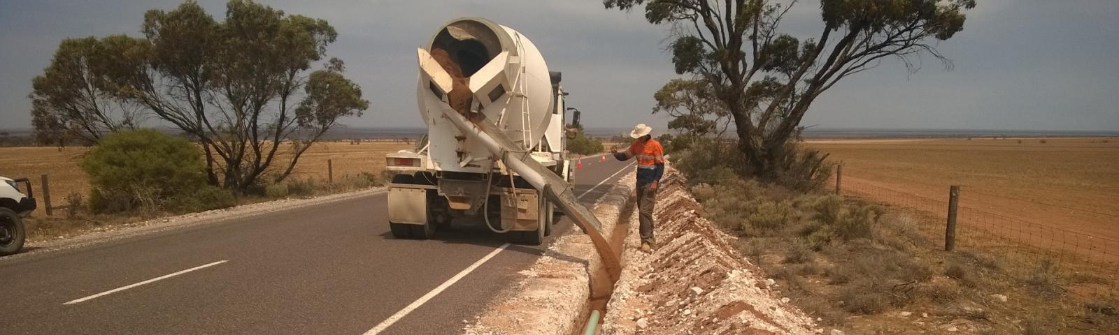 Laying pipe at Coolanie Water Scheme in South Australia. A concrete truck and a worker are covering a light blue pipe with material in a trench to the right side of a country road.