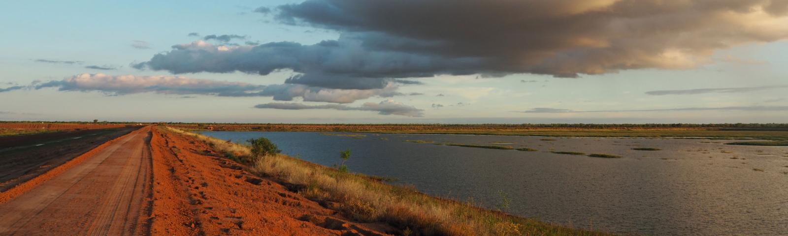 A body of water is shown beside a red dirt road, a row of grass runs along the water’s edge separating the water from the dirt road.