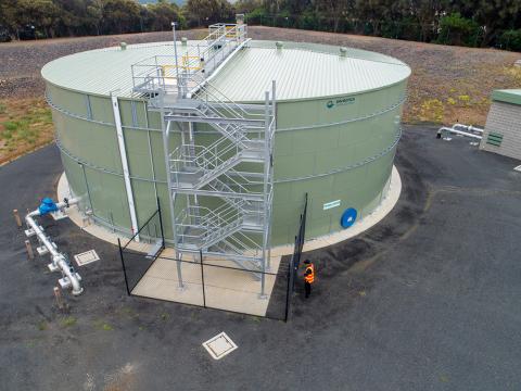 An aerial photograph of the drinking water tank at Wimbledon Heights on Phillip Island. There is a large, circular, green tank surrounded by black asphalt. There is a silver staircase attached to the tank which provides access to the top of the tank