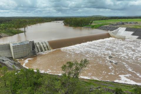 Significant volumes of water flowing from the Fitzroy River in the background, spilling over the completed Rookwood Weir and creating a whitewash effect as the water continues to flow down river from the weir. The river is surrounded by green pastural land.