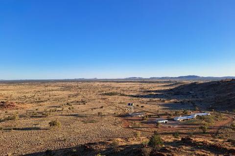 An image of a small township in a remote area. There is a cluster of small light-coloured buildings and an elevated water tank in the foreground, surrounded by a large expanse of arid bushland with red dirt and rocks, grasses, trees and a bright blue sky.