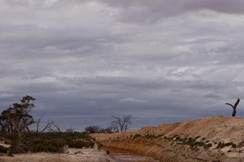 A muddy field with a floodwater river running through with storm clouds above.