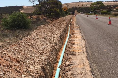 Laying pipe at Coolanie Water Scheme in South Australia. The light blue pipe is laid in a trench in the centre of the image, to the left of a country road.