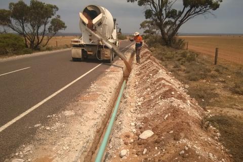 Laying pipe at Coolanie Water Scheme in South Australia. A concrete truck and a worker are covering a light blue pipe with material in a trench to the right side of a country road.