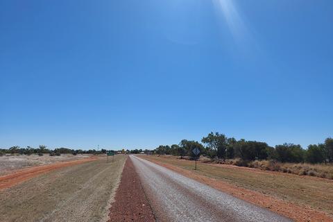 Single-lane road in the outback leading into the Dajarra community, with a blue sky. The road is surrounded by grass, red dirt and scrubby vegetation.