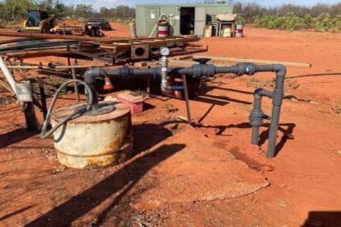A bore sitting on an aged concrete pad with grey pipes running between the top of the bore and the ground beside it. A green shed and tractor sits in the background with scrubby bush behind them. Everything is sitting on and surrounded by red dirt.