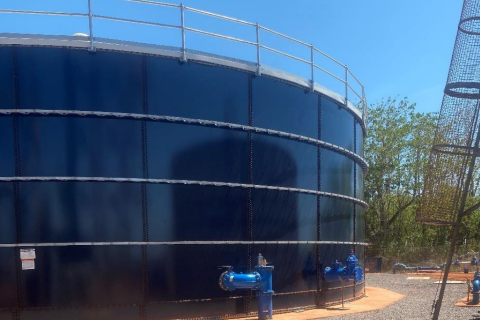 Photo of a large dark blue water tank in Galiwin’ku. There are blue valves at the bottom and a silver metal fence on top. There is pavement in the foreground and blue sky and green vegetation in the background. 