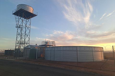 Photograph of a water treatment plant, including a water tower and circular water storage tank behind a barbed wire fence. 