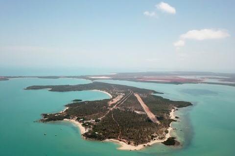 An aerial view of Gunyangara on the Gove Peninsula in north-east Arnhem Land. There is blue water and a coral reef surrounding a vegetated island. The image shows an airport runway and some roads, buildings and boats.