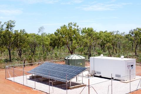 An example of a mini plant compound. A concrete pad surrounded by fencing with a water storage tank, solar panels and shipping container that houses water treatment equipment. There is red dirt surrounding the compound and native trees behind the compound. 