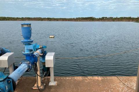 Dithery Creek Dam with dark blue water and bright blue dam equipment, including a water pump and pipes. There is a concrete dock in the foreground where the equipment sits. A blue cloudy sky and green vegetation is in the background on the horizon.