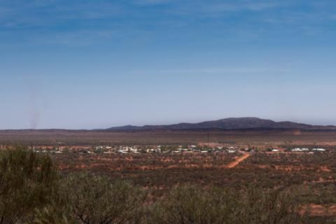 Looking across a desert landscape in the Northern Territory, with native trees and bushes in the foreground and a remote community in the distance. 