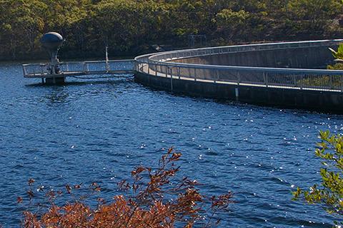 Large dam with short dam walls on the right and bushland in the background, sunny day.