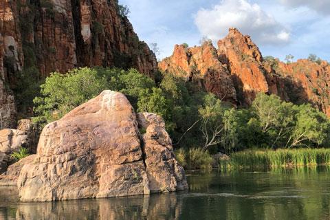 Lake in foreground with rock formations, cliffs and green shrubs behind.