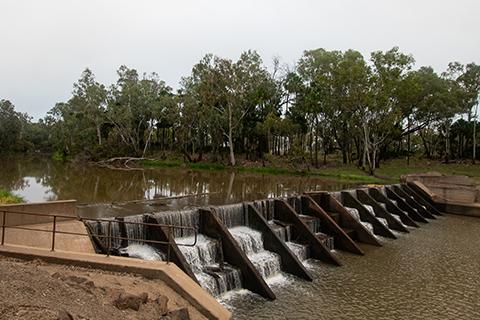 River with a weir across it, trees in the background and dirt in the foreground. 