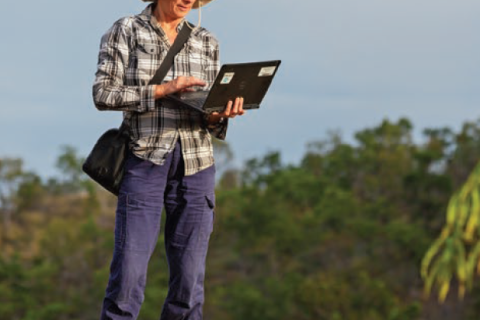 Photograph of a researcher standing, wearing a hat and holding a laptop.