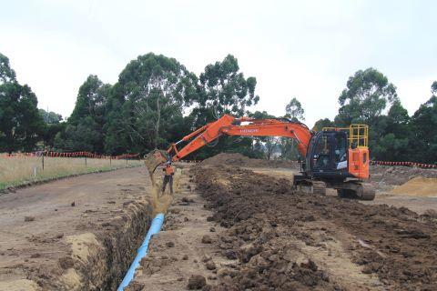 Pipeline being installed in a trench by machinery, surrounded by dug up soil with a tree line in the background. 