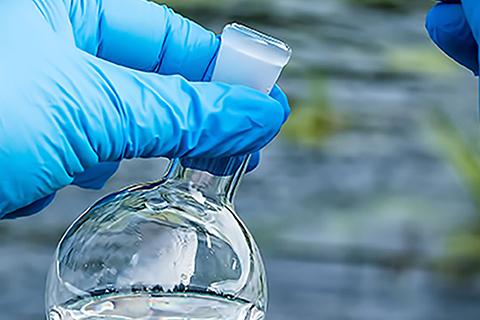 Close up of hands wearing blue gloves holding a small beaker with a water sample, and a river in the background.