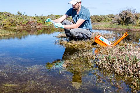 A male scientist kneels near water, using collection equipment.