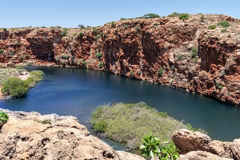 Blue waterhole surrounded by red rocky cliffs and sparse green vegetation under a clear sky.