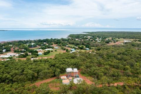 An aerial view of a small community. There are water tanks in the foreground, with a band of trees and scattered buildings behind them, leading to water. 