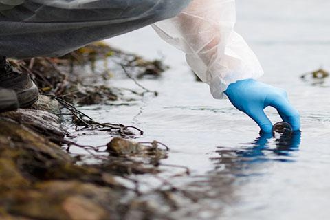 Person in protective clothing and blue gloves gathering a water sample from the rocky bank of a body of water.