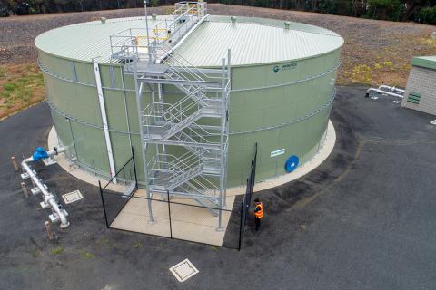An aerial photograph of the drinking water tank at Wimbledon Heights on Phillip Island. There is a large, circular, green tank surrounded by black asphalt. There is a silver staircase attached to the tank which provides access to the top of the tank